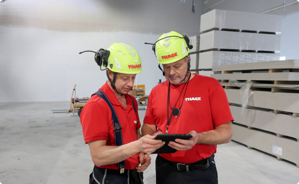 Construction workers using Dalux Field software on a tablet to review project plans in an indoor construction site, wearing safety helmets.